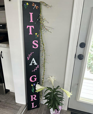 Gender reveal sign with 'It's a Girl' birth announcement in pink letters and floral decorations, stenciled on a large chalkboard using magnetic chalkboard stencils and painting pens, displayed with a potted plant