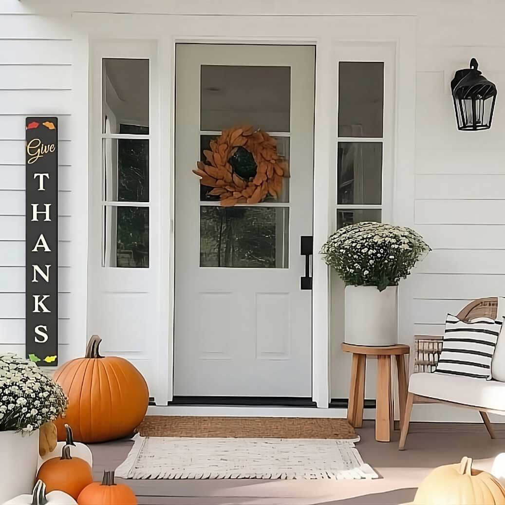 A large outdoor chalkboard sign with fall chalk art stands next to front door of a home with fall decorations, the large chalkboard is stenciled ‘Give Thanks’ by using Plata magnetic ‘Give’ word stencil, letter stencils, fall leaf stencils and colored with erasable paint pens to craft a large fall welcome sign