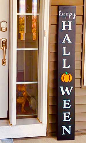 Black vertical chalkboard sign standing by a front door with 'HAPPY HALLOWEEN' in white and orange stenciled letters, enhanced with a pumpkin design and made using magnetic letter stencils and chalk markers from a DIY porch sign kit, with a dog peering from behind the glass door.