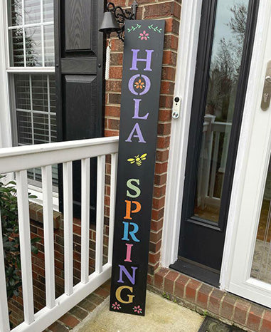Tall outdoor chalkboard sign next to a front door stenciled 'HOLA SPRING' in colorful letters with flowers and a bee, crafted using Plata Porch Chalkboard and Magnetic Stencil kit and Spring Stencils to create a spring welcome sign