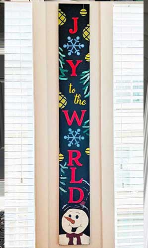 Tall chalkboard sign hanging between window shutters, with 'JOY to the WORLD' in colorful letters, snowflake accents and a smiling snowman at the bottom, created with chalkboard stencils to create a Christmas sign