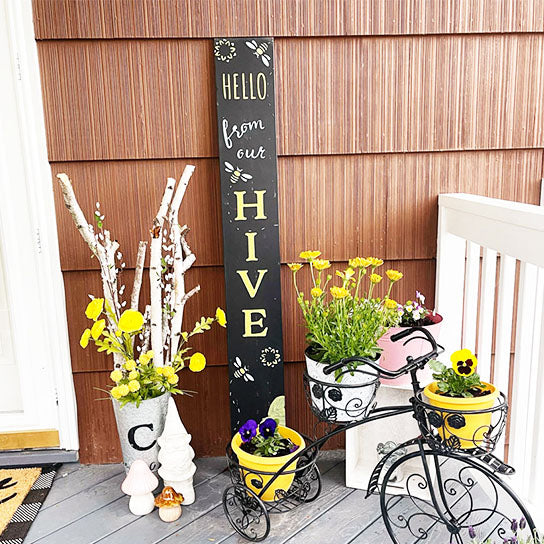 A large outdoor chalkboard stenciled 'hello from our hive' with sunflowers and bees next to a front door  to create  a bee welcome vertical sign