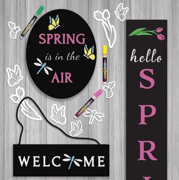 A collection of spring chalkboards surrounded with spring stencil set and colorful chalk markers- &#39;Spring is in the Air&#39; on oval chalkboard with yellow and blue butterflies and a dragonfly, a hanging door welcome sign with a dragonfly for the letter o and a large outdoor chalk board &#39;hello spring&#39; with pink tulips 