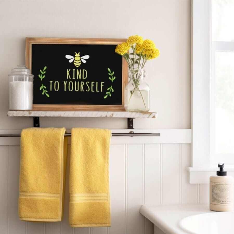 a pine framed rustic chalkboard with inspirational chalk art stenciled with bee chalk art and chalkboard lettering to say &#39;BEE kind to yourself&#39;, chalkboard stands on shelf in bathroom next to a candle and vase of yellow flowers