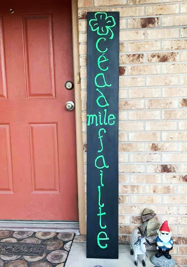 Rustic vertical welcome sign placed beside a red door, featuring the Irish greeting 'Céad Míle Fáilte' in elegant green script above a shamrock, inviting warmth and hospitality for St. Patrick's Day