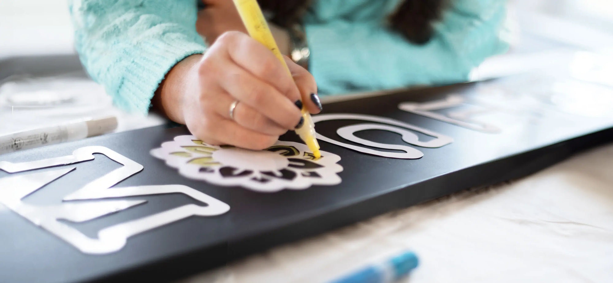 Close up shot of a woman coloring a sunflower stencil with a yellow chalk marker on a large outdoor chalkboard welcome sign 