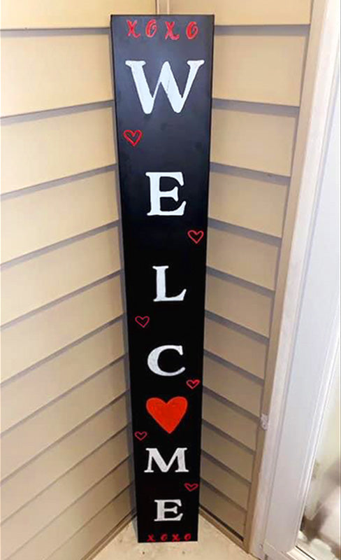 A customer photo of a DIY Valentine's Day Welcome Sign crafted on our 5 foot porch chalkboard using magnetic chalkboard letter stencils to spell welcome painted white. The O in welcome is replaced with a red stenciled heart and the top of the sign uses x and o stencils for decoration.