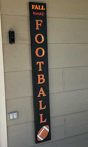 Outdoor blackboard sign saying 'FALL means FOOTBALL' stenciled in bold orange letters with a white football stencil, placed beside a house door crafted with DIY sign kit to create a football themed welcome sign