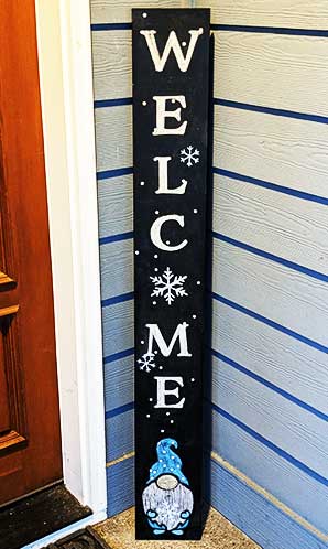 Vertical outdoor chalkboard sign placed beside a blue siding door with 'WELCOME' stenciled in bold, white letters, adorned with snowflake designs and a blue gnome at the bottom crafted with gnome sign stencils, handcrafted porch welcome sign 