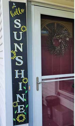 Tall black magnetic chalkboard sign beside a red door, stenciled 'hello SUNSHINE' in white letters with bright yellow sunflowers invoking a cheerful, sunny welcome, created with chalkboard paint letter stencils and sunflower stencils to create a DIY summer welcome sign