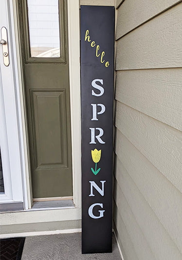A creative DIY outdoor chalkboard sign leaning against a house siding beside a green door, cheerfully announcing "hello SPRING", crafted with magnetic stencil letters and a tulip for the 'I' in spring to create a vertical spring welcome sign 