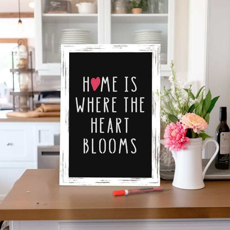 a chalkboard with a rustic white farmhouse frame standing on a kitchen counter next to a pitcher of flowers, the chalkboard is stenciled with chalk art &#39;home is where the heart blooms&quot; 
