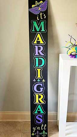 Vibrant Mardi Gras chalkboard sign standing indoors, featuring green, yellow and purple stenciled letters spelling 'it's MARDI GRAS y'all' with a festive mask and stars to create a colorful Mardi Gras chalkboard sign