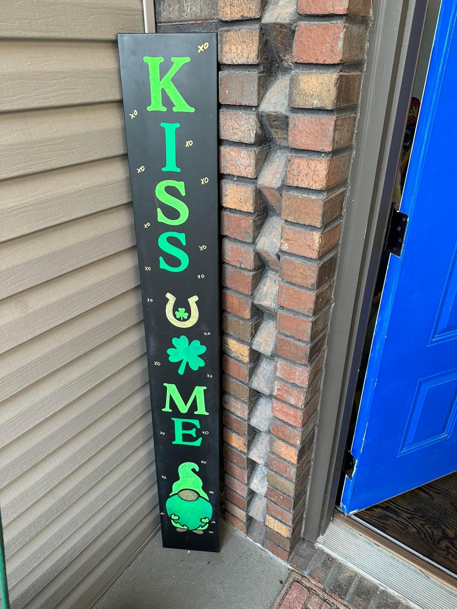 Tall vertical outdoor chalkboard sign stenciled 'KISS ME' in large green letters with a cute gnome at the bottom created by using gnome stencil kit,, surrounded by small white stars and shamrocks, for a whimsical St. Patrick's Day welcome sign