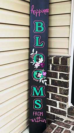 Tall outdoor blackboard sign leaning against a house corner with 'happiness BLOOMS from within' stenciled in vibrant pink and green letters, accented with a blossoming flowers, created using a DIY chalkboard sign kit for an inspiring home sign