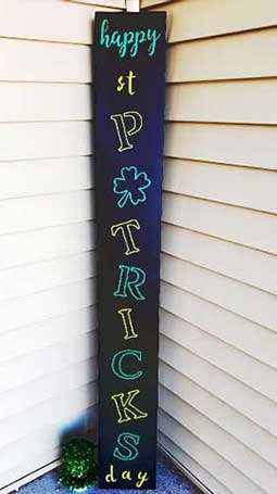 Vertical outdoor chalkboard sign placed against house siding with 'happy st PATRICKS day' in green and white stencil letters, adorned with shamrock designs, handcrafted with chalk markers for fun outdoor St. Patrick's Day decoration