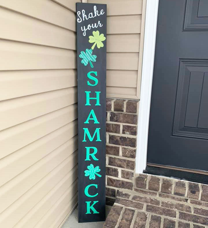 Vertical metal welcome sign porch chalkboard placed by a home's entryway, stenciled 'Shake your SHAMROCK' in bold green and white letters with shamrock accents with letter stencils and chalk markers, crafted for St. Patrick's Day decorations
