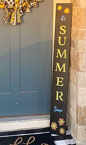 Tall, vertical large chalkboard sign by a blue front door reading 'it's SUMMER time' in yellow and white stenciled letters, accented with sunflower designs, made with a DIY outdoor signs kit using magnetic chalkboard stencils and erasable paint markers for a seasonal welcome