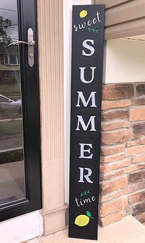 Tall, vertical large chalkboard sign by a blue front door reading 'it's SUMMER time' in yellow and white stenciled letters, accented with sunflower designs, made with a DIY outdoor signs kit using magnetic chalkboard stencils and erasable paint markers for a seasonal welcome