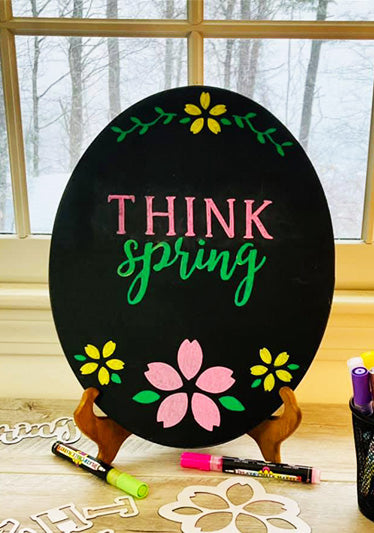 Oval spring chalkboard sign stenciled 'Think Spring' in pink and green letters using capital letter stencils and cursive stencils with yellow and pink flowers surrounding the words crafted with flower stencils