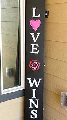 Vertical outdoor blackboard sign against a yellow wall with 'LOVE WINS' in bold, white letters, featuring a pink heart and a red rose design, crafted with a DIY sign craft kit using magnetic stencils and chalk paint markers to make a Valentines Day Door Sign