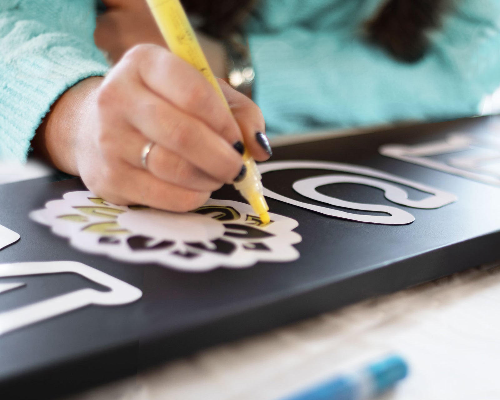 Close up shot of a woman coloring a sunflower stencil with a yellow chalk marker on a large outdoor chalkboard welcome sign 