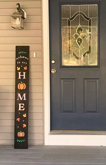 "Welcome to Our Home" fall front porch DIY sign by painting letter stencils on a vertical sign chalkboard and decorated with pumpkin and fall leaf chalk art