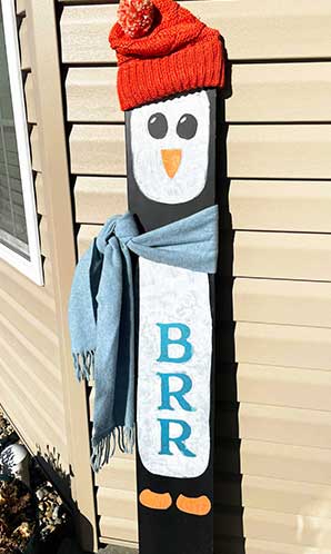 Decorative outdoor blackboard sign colored to create a penguin with 'BRR' written in blue letters, topped with an orange winter hat and blue scarf, assembled using a craft kit with magnetic stencils and chalk markers, adding a playful touch to outdoor winter decor.