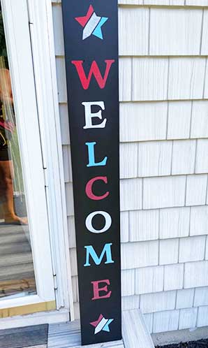 Large chalkboard sign placed against a house siding, spelling out 'WELCOME' in red, white and blue painted letters flanked by patriotic star designs, crafted with a DIY sign kit to create a vibrant 4th of July greeting sign