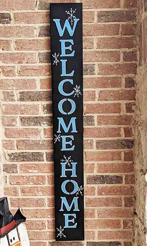 Handmade outdoor blackboard sign mounted on a brick wall with 'WELCOME HOME' in large, white stenciled letters, embellished with snowflake accents, created using a DIY sign kit with magnetic chalkboard stencils and chalk markers for a winter welcome sign
