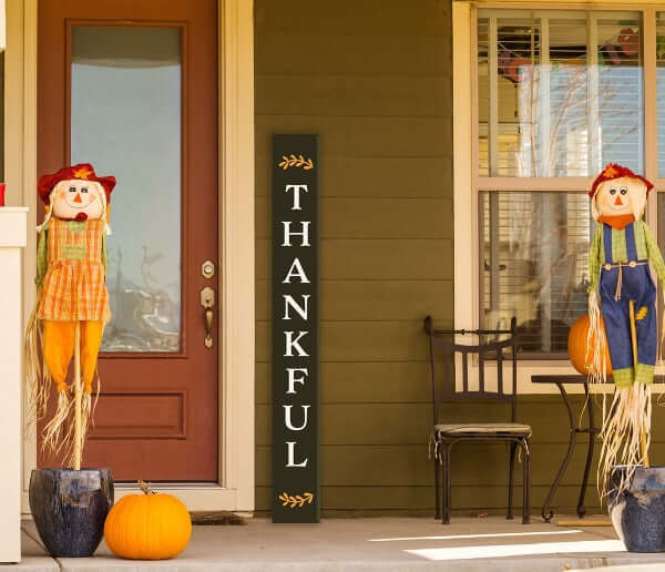A Plata porch chalkboard stenciled to craft a Thankful Tall Door Sign next to a front door of a colonial home decorated for autumn with scarecrows
