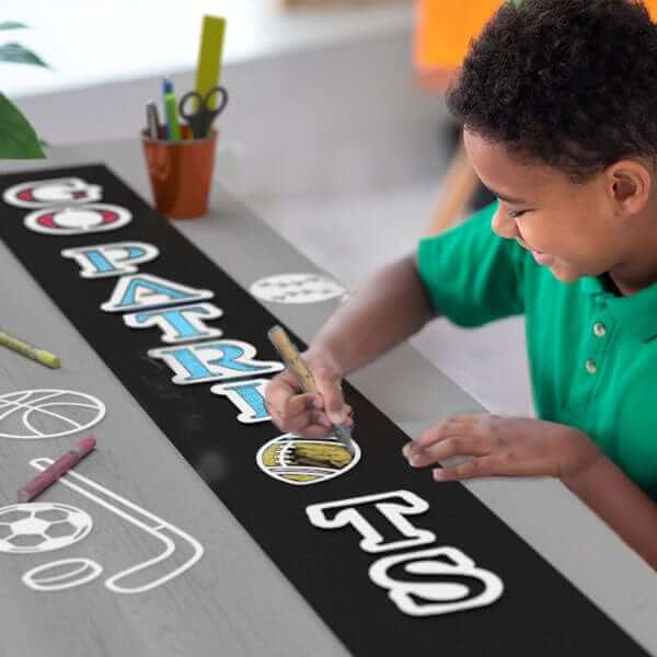 Boy crafting a sports sign &quot;Go Patriots&quot; football sign with letter stencils and painting pens on a large chalkboard sign