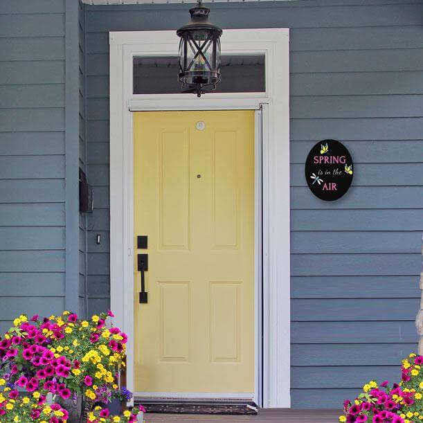 An finished spring craft project- an oval spring chalkboard sign hung next to the front door of a modern farmhouse. Spring is in the Air Sign craft with chalkboard markers and spring stencils for chalkboards by Plata Chalkboards 